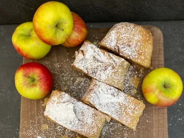 Apfelstrudel liegt auf einem Holzbrett, umgeben von Äpfeln.
