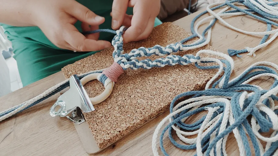 Hands weaving macramé on cork board.