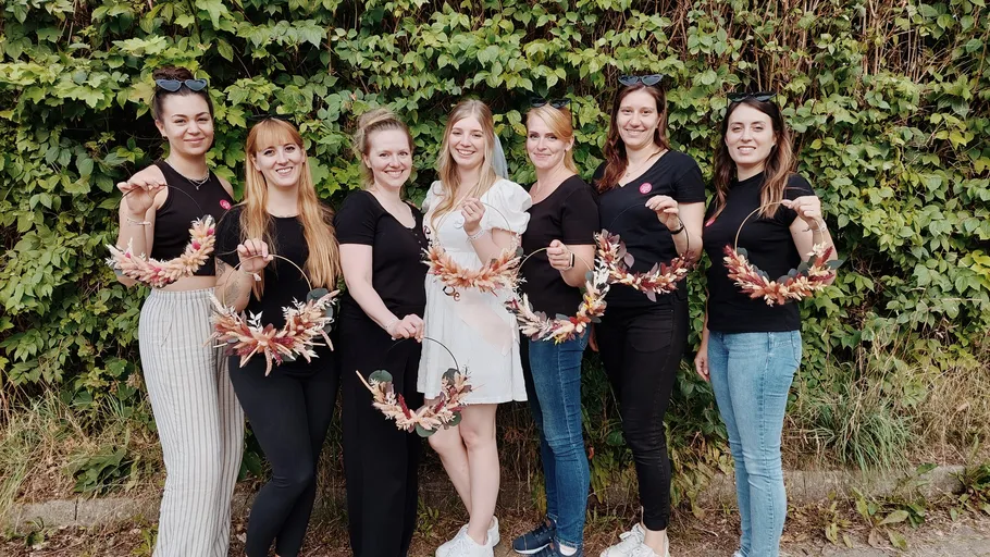 Seven women holding floral wreaths, leafy background.
