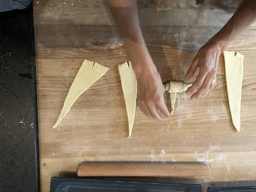 Pieces of dough are formed into croissants on a wooden table.