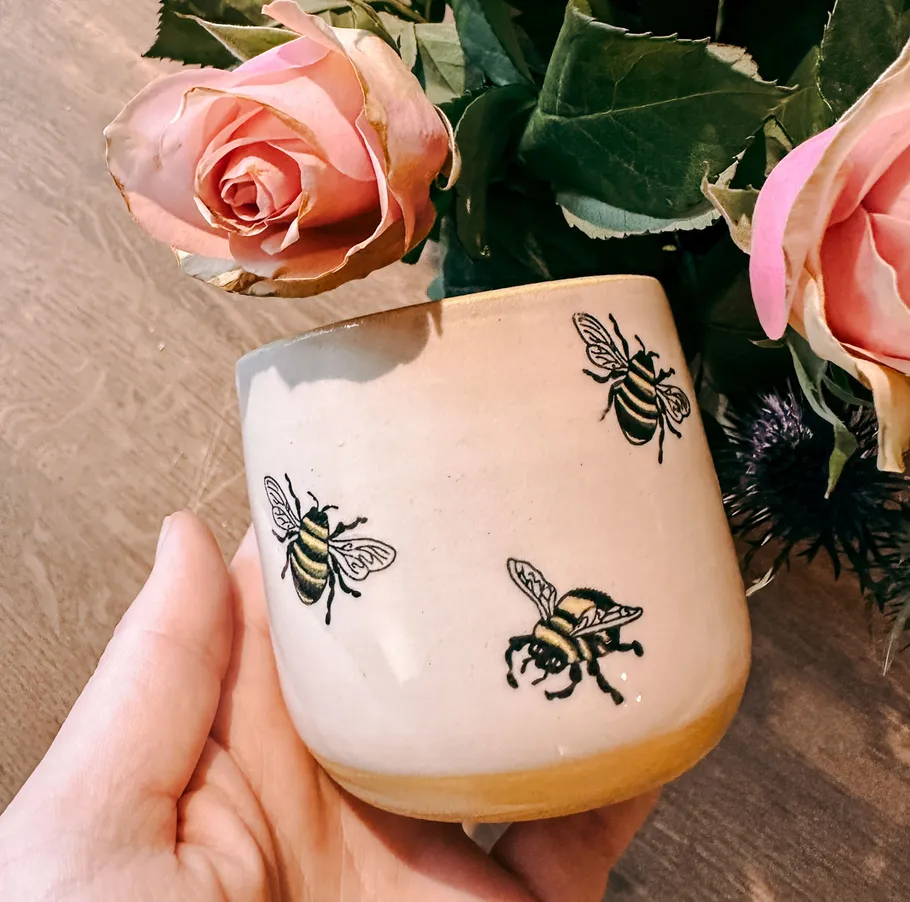 Hand holding bee-decorated ceramic mug near roses.
