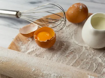 Eier, Schneebesen und Mehl auf einer Holzplatte.