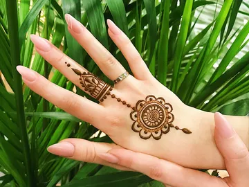 Hände mit Henna-Muster vor grünen Pflanzen im Hintergrund.