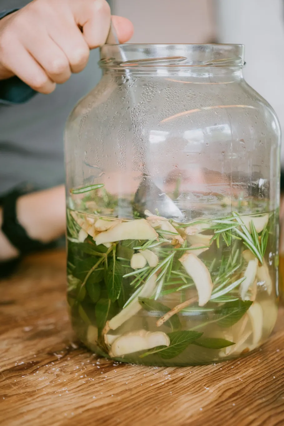Hand stirring herbs in large glass jar.