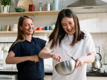 Zwei Frauen lachen und backen in einer modernen Küche.