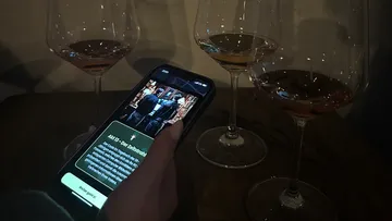 Hand hält Telefon neben drei Weingläsern.