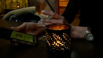 Hand hält Getränk, Handy, Kerze auf Tisch.