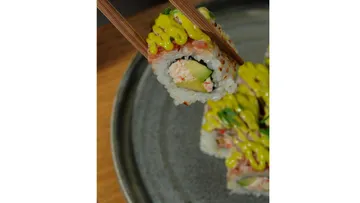 Sushi roll held by chopsticks over plate.
