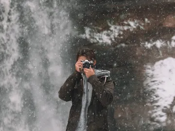 Man with camera takes pictures in front of a waterfall in the snow.