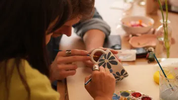 Two people painting a floral mug indoors.