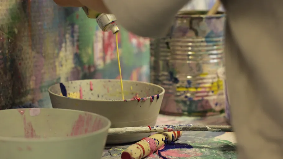 Person gießt Farbe in Schüsseln auf unordentlichem Tisch.