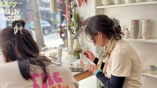 Two women seated, crafting near pottery display.