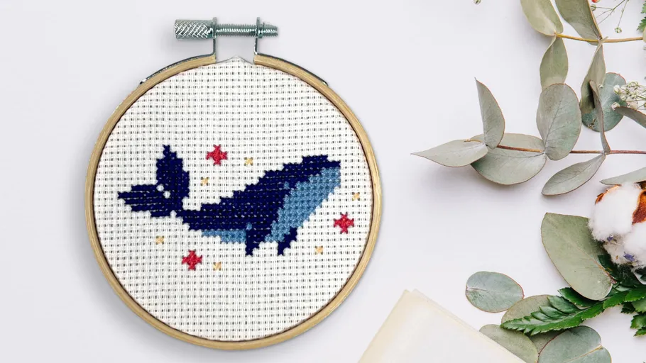 Whale cross-stitch in wooden hoop, surrounded by leaves.