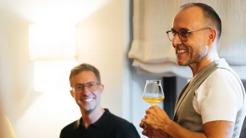 Two men smiling, one holding a drink indoors.