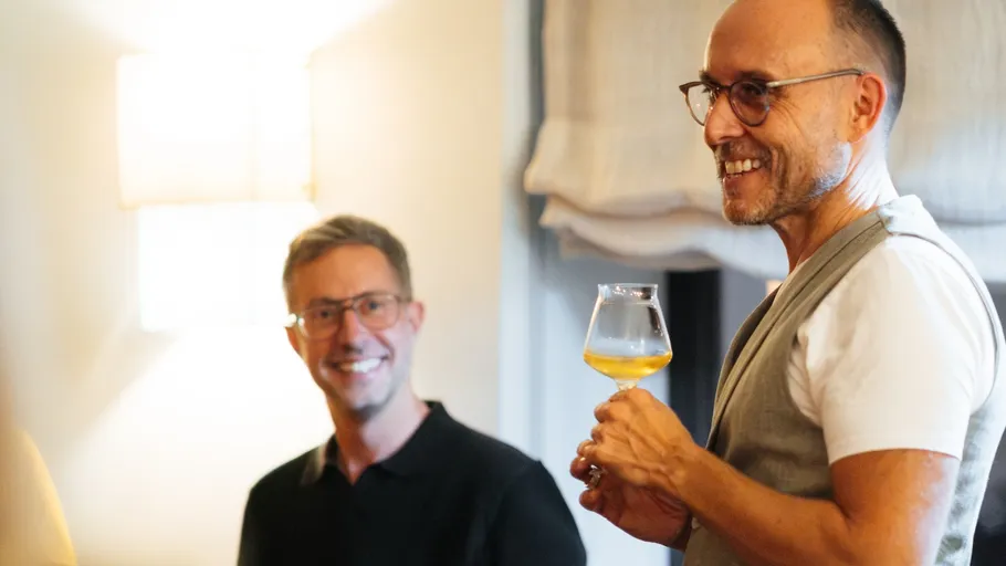 Zwei lächelnde Männer, einer hält ein Glas drinnen.