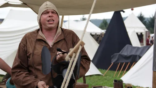 Person in medieval clothing, holding helmet, tents behind.