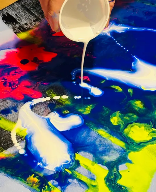 Hand gießt weiße Farbe auf bunte Leinwand.