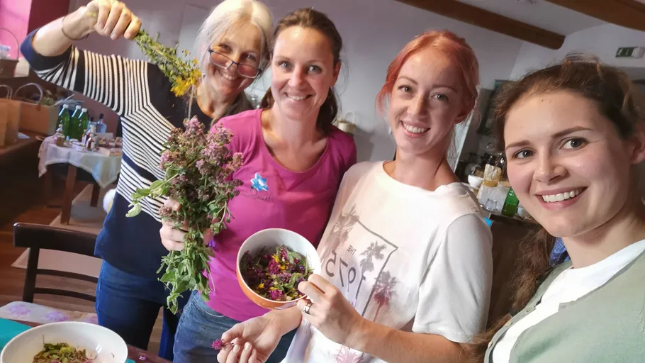 Four women smiling with herbs and bowls indoors.