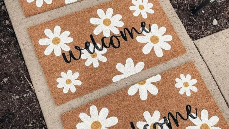 Three welcome mats with daisy design on pavement.