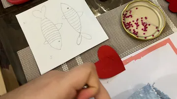 Hand nahe Fischzeichnung, Herz, Perlen auf Tisch.