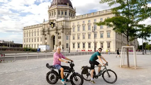 Two people cycling in front of grand building.