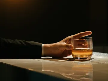 A hand grabs a glass of whiskey on a table.