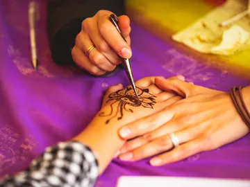 Hands get an ornate henna pattern on purple table.