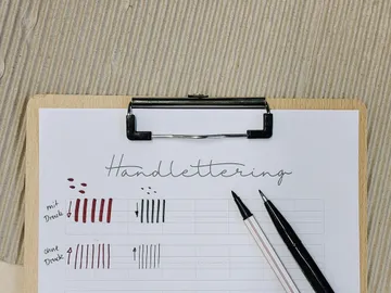 Das Objekt ist ein Schreibblock mit Handlettering. Zwei Stifte liegen darauf.
