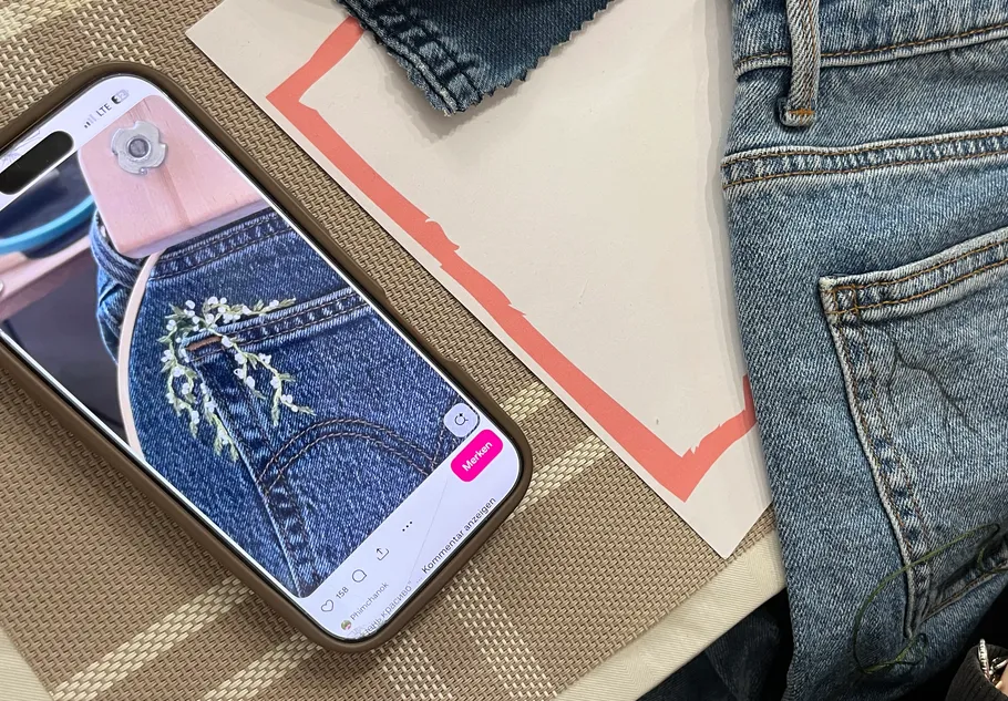 Handy zeigt bestickte Jeans neben Denimstoff.