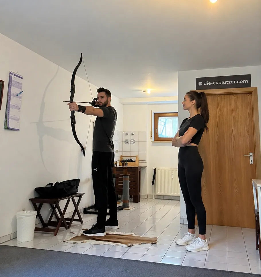Man practices archery while woman observes indoors.