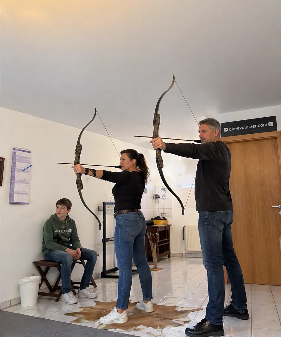 Two adults practice archery indoors, teenager watching.