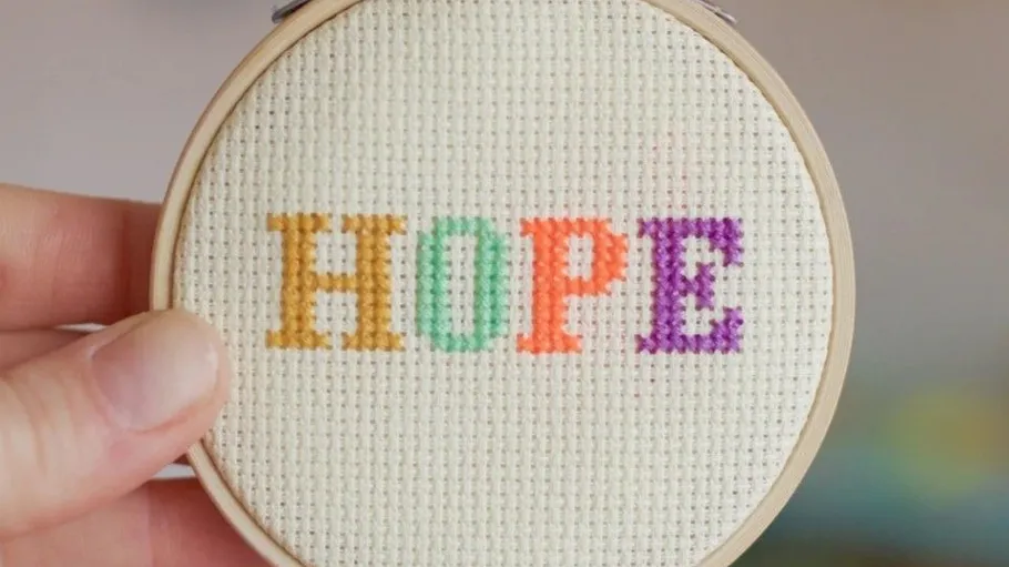Hand holding cross-stitched hoop with 'HOPE'.