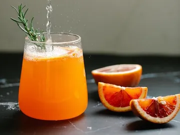Orange juice is poured into a glass surrounded by oranges.