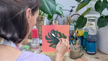 Woman painting leaf, surrounded by plants and paints.