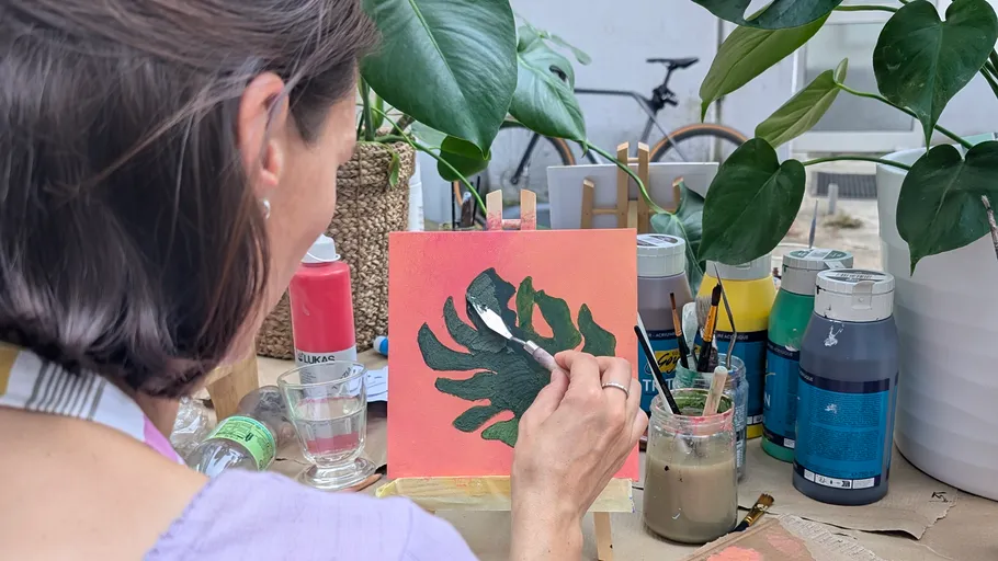 Woman painting leaf, surrounded by plants and paints.