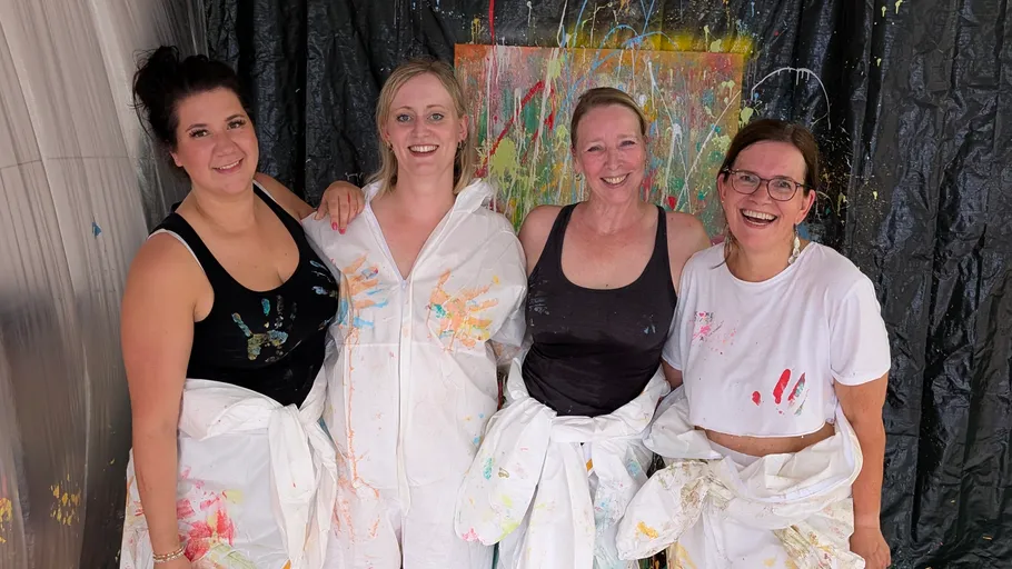 Four women in paint-splattered clothes smiling indoors.