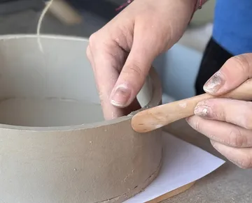 Hands shaping clay bowl with tool.