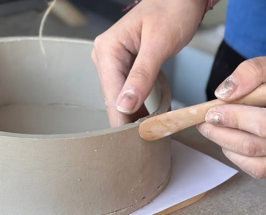 Hands shaping clay bowl with tool.