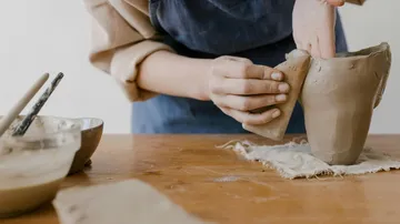 Person shaping clay pottery on wooden table.