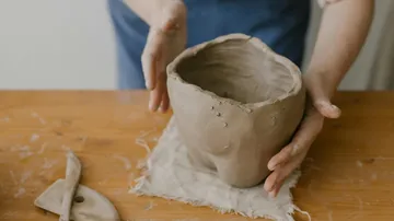 Person shaping clay pot on wooden table.