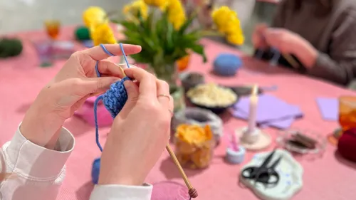 Hands knitting with blue yarn on pink table.