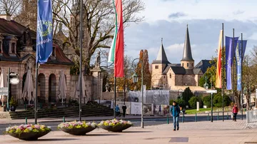 Street with flags, person walking, church towers visible.
