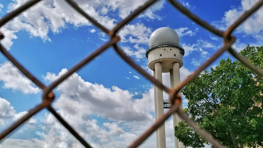 Radarturm hinter Maschendrahtzaun, blauer Himmel.