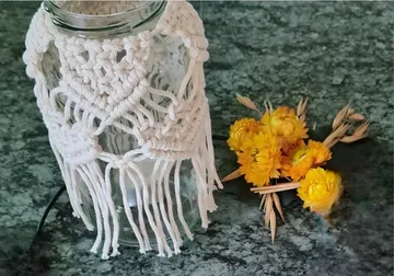 Macramé-covered jar beside yellow flowers on table.