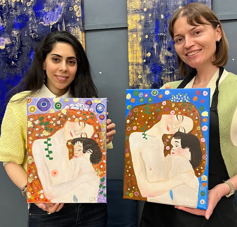 Two women holding colorful paintings in art studio.