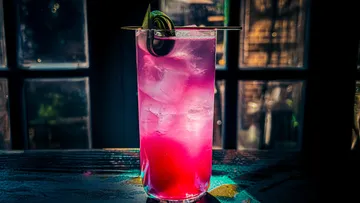 Pinkfarbener Cocktail mit Eis und Olive, schummrige Bar.