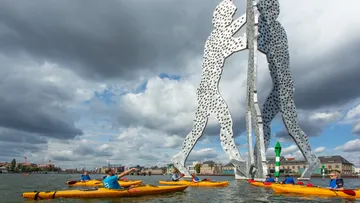 Kajakfahrer paddeln nahe Metallskulptur auf Fluss.