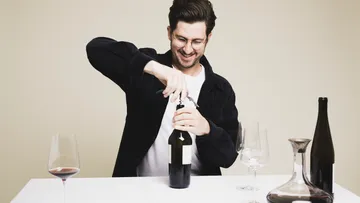 Mann öffnet Weinflasche am Tisch mit Gläsern.