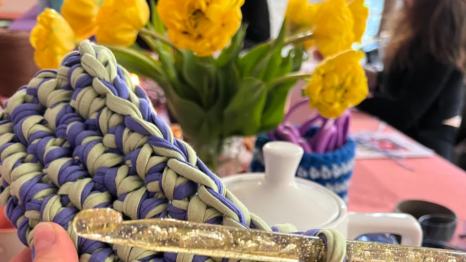 Hand mit Häkelarbeit und gelben Blumen im Hintergrund.
