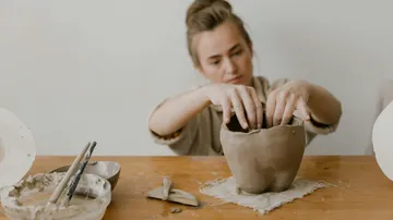 Person shaping clay pottery on wooden table.